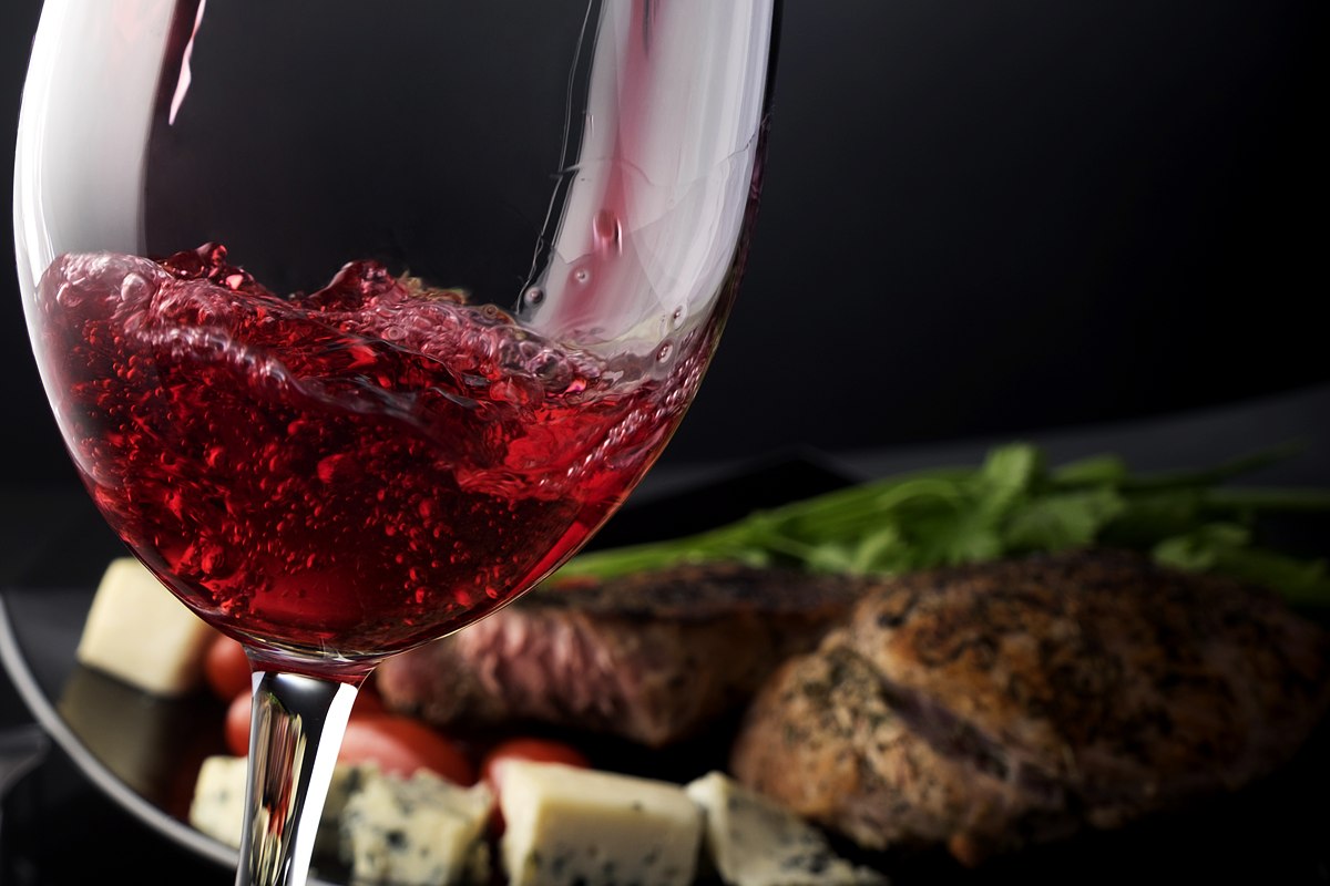 Rotwein wird in ein Glas geschenkt, dahinter Steak, Kirschtomaten und Käse